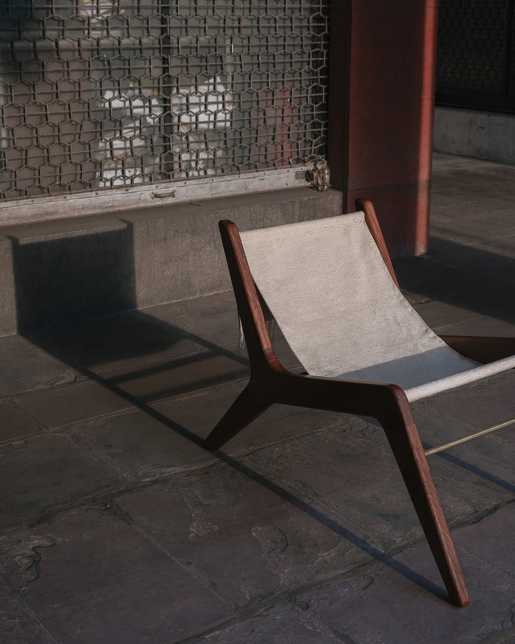 A mid-century modern wooden lounge chair with a light beige canvas sling seat, positioned on a stone floor in front of a metal screen.